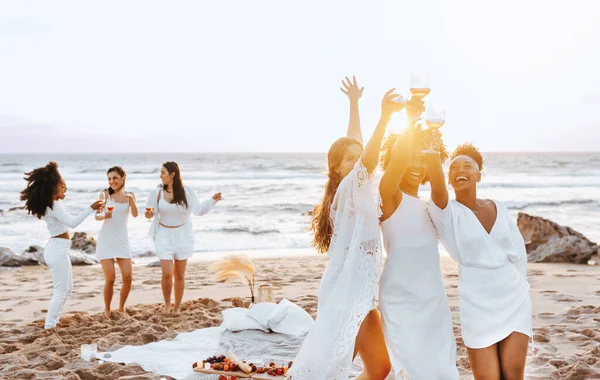 Carefree ladies bride team toasting wine glasses while having hen party and dancing on the beach at coastline. Excited female friends enjoying their vacation