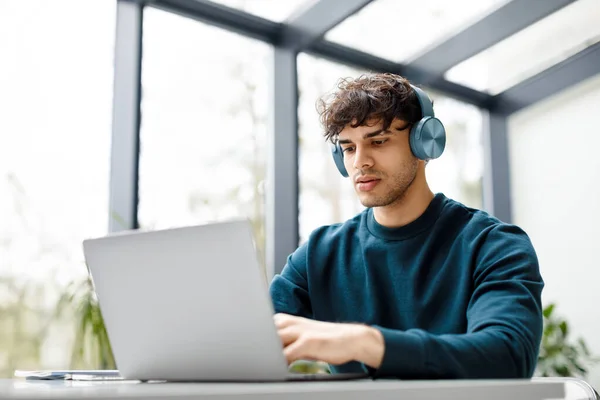 若いヨーロッパの男性デザイナーは 現代のガラスオフィスのインテリアでノートパソコンで作業しながらヘッドフォンで音楽を聞いています コピースペース フリーランス オンライン求人コンセプト — ストック写真