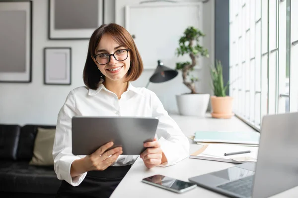 快乐的女经理穿着正式服装 戴着眼镜 在舒适的办公室内部用平板电脑 对着相机微笑 自由自在的空间 具有设备 工作演示 博客和学习功能的现代商务应用程序 — 图库照片