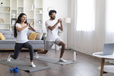 Evde antrenman. Spor yapan Hintli çift laptopun önünde kalça egzersizi yapıyor, mutlu genç doğu eşleri online dersler izliyor ve oturma odasında egzersiz yapıyor, birlikte spor yapıyorlar.