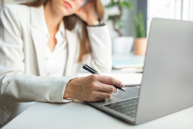Resmi görevdeki düşünceli Avrupalı iş kadını elinde kalem, ofis içi dizüstü bilgisayarın önünde oturuyor, seçici odaklanma, yakın çekim. Uzaktan buluşma, iş, video görüşmesi, alet üzerinde iş.