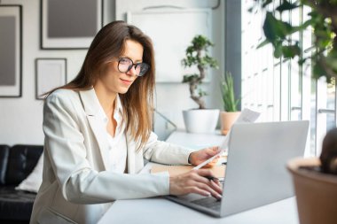 Ofiste evrak ve dizüstü bilgisayarla çalışan pozitif iş kadını, masa başında oturan, bilgisayar kullanan ve belgeleri kontrol eden, uzaktan çalışmanın keyfini çıkaran, serbest alan