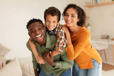 Aile Mutluluğu. Neşeli farklı ebeveynler ve kameraya gülümseyen küçük oğulları modern iç mekanda poz veriyorlar. Mutlu baba, anne ve çocuğunun portresi.