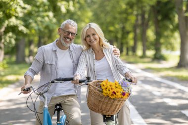 Yetişkin eşlerin parkta bisiklet sürerken kamerada poz vermesi, eski bisikletlerin üzerinde oturan, kucaklaşan ve gülümseyen, aktif yaşam tarzını ve açık havada eğlenen mutlu bir çiftin portresi.