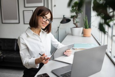 Dijital tablet ve dizüstü bilgisayarla çalışan, rahat bir ofis veya kafede masa başında oturan, iş için modern teknolojileri kullanan, ağ kurmaktan keyif alan genç bir iş kadını.