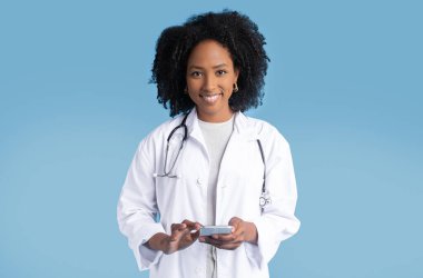Beyaz önlüklü mutlu siyah bayan doktor telefon kullanır, sohbet eder, mavi stüdyoda izole olur. Tıp ve sağlık hizmetleri, uzaktan çalışma uygulaması, profesyonel danışma