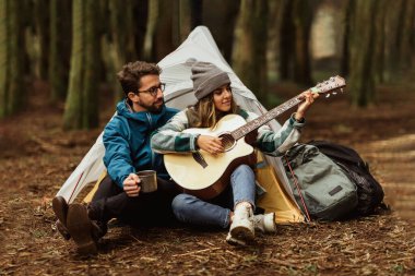 Ormandaki ceketli mutlu beyaz çift çadırı olan, gitar çalan, müzik dersi alan, açık havada gezinin tadını çıkarıyor. Aktif yaşam tarzı, macera ve turizm, kampla yürüyüş