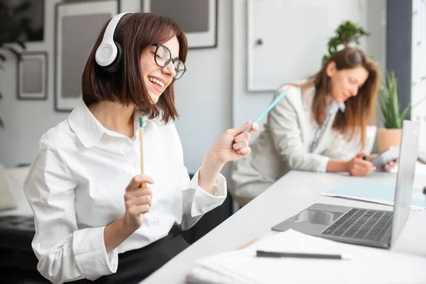 Excited young lady office worker in headphones enjoying music and imagining playing the drums while working on laptop in office, sitting with colleague at workplace