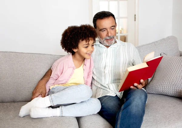 和爷爷一起舒服地看书 快乐的西班牙裔孙子和他的祖父一起读书 周末享受传统的闲暇 坐在家里现代客厅的沙发上 — 图库照片
