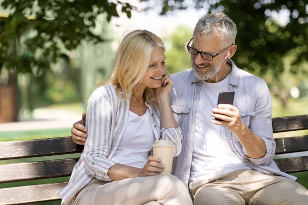 Romantic Older Couple With Smartphone Relaxing On Bench In Summer Park, Happy Mature Man And Woman Listening Music Together, Sharing Earphones And Drinking Takeaway Coffee, Enjoying Outdoor Leisure