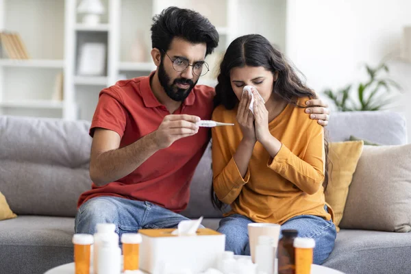 Portrait Of Worried Indian Man Taking Care Of His Ill Wife At Home, Caring Husband Checking Temperature And Looking At Thermometer With Shock While Sick Woman Blowing Nose Into Napkin, Free Space