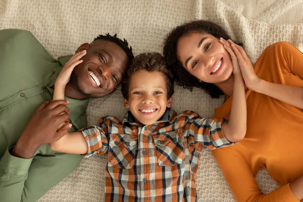 Portrait Of Happy Diverse Family Of Three Indoors, Cheerful Multicultural Parents And Their Little Son Lying And Smiling To Camera, Top View. Kid Boy Hugging His Mommy And Daddy