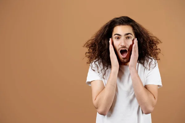 Omg. Emotional Shocked Guy With Curly Long Hair Touching Face, Expressing Amazement Looking At Camera With Open Mouth Over Beige Background In Studio. Portrait Of Surprised Amazed Young Man