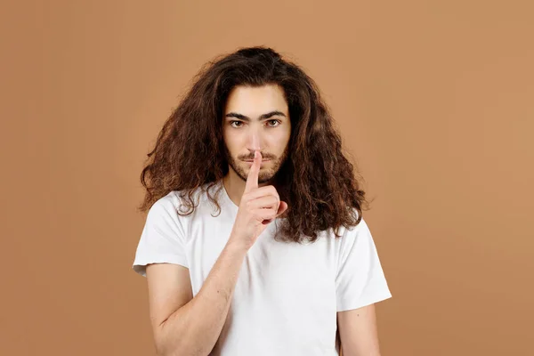 Be Silent. Latin Guy With Long Hair Making Zip Your Lip, Finger on Lips Gesture, Encouraging Silence and Secrets Over Brown Studio Background. Portrait Of Young Man Showing Hush Sign