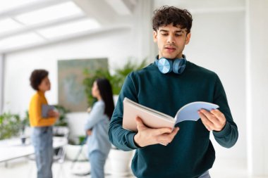 Ciddi üniversite öğrencisi elinde kitapla ders ya da sınava hazırlanıyor, arka planda bayan sınıf arkadaşlarıyla modern üniversite seyircilerinin arasında duruyor.