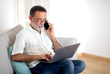 Laptop kullanırken cep telefonuyla konuşan, iletişim kuran ve evde koltukta oturan, gözlük takan İspanyol kıdemli bir adam. Olgun İş Adamı Cep Telefonu Konuşması Yapıyor