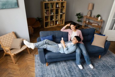 Rahat Japon genç çift evdeki modern oturma odasında, yüksek açılı koltukta dinlenirken akıllı telefon kullanıyor. Asyalı Eşler Mobil Uygulama ile Mesajlaşıyor ve Sosyal Medya İçeriğini Birlikte Paylaşıyorlar