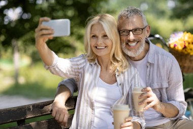 Mutlu, olgun bir çift parkta bankta otururken selfie çekiyor, neşeli yaşlı eşler akıllı telefonlarıyla fotoğraf çekiyor, paket kahve içiyor, dışarıda pikniğin tadını çıkarıyor,