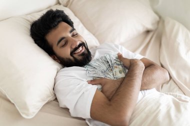 Yatakta uyurken dolar tutan gülümseyen Hintli genç adam, sakin bir milenyum doğusundaki adam yatak odasında dinlenirken bir sürü banknota sarılıyor. Para tasarrufu konsepti, yakın çekim