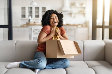 Heyecanlı genç Afrikalı Amerikalı Kadın Evde Yeni Giysilerle Kutuyu Açma, Mutlu Siyah Kadın Paket Teslimatı 'ndan sonra tişörtünü çıkarıp kucaklıyor, Çevrimiçi alışveriş ile tatmin oluyor,