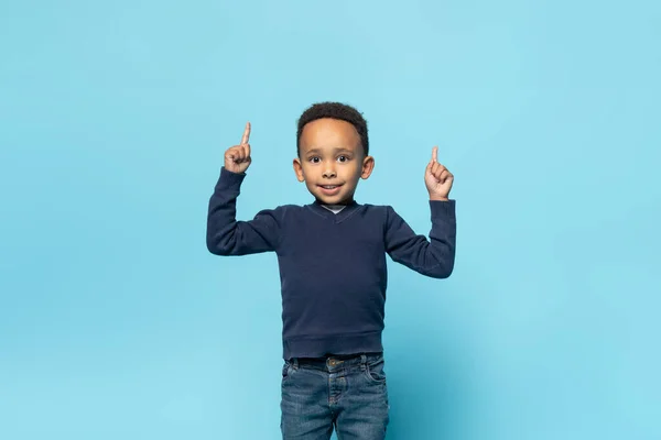 看看这个高兴的黑人小男孩用双手指着空旷的空间 微笑着 孩子们展示广告的地方 站在蓝色的工作室背景上 — 图库照片