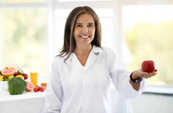 Cheerful adult caucasian nutritionist doctor in white coat shows red apple in clinic office interior. Weight loss and health care recommendation, advice by professional, body care