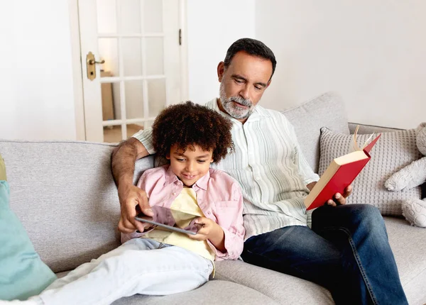 多代家庭休闲 外公在看他爷爷的纸质书 坐在沙发上在家里 的时候 拉美裔孙子用的是平板电脑 数字和传统的周末业余时间概念 — 图库照片