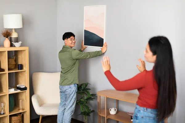 Asian Millennial Spouses Hanging Poster In Frame On Wall Together Standing At Modern Living Room In Home Interior, Decorating House With Pictures After Renovation. Selective Focus