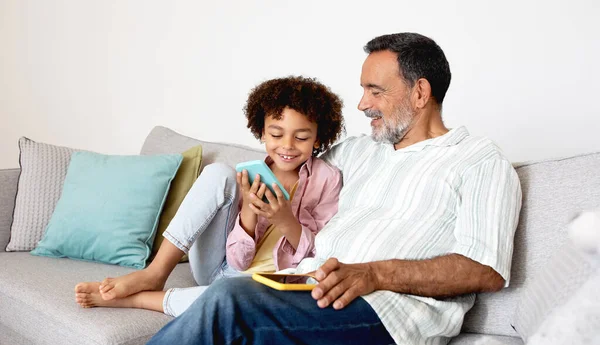 世世代代的科技乐趣 快乐的祖父和孙子用他们的智能手机 一起上网浏览和移动游戏 坐在沙发上 把手机放在家里 — 图库照片