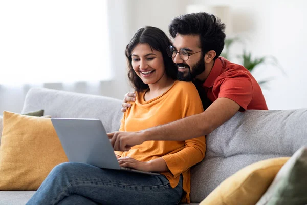 Great Website. Happy Young Indian Couple Using Laptop Together At Home, Cheerful Eastern Spouses Booking Vacation Online Or Shopping In Internet While Resting On Sofa, Man Pointing At Computer Screen