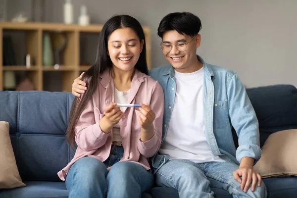 Fertility And Childbirth. Joyful Asian Couple Holding Positive Pregnancy Test Sitting On Sofa At Home. Happy Family Dreaming Together Awaiting Baby Concept. Shot Of Parents-To-Be With Good News