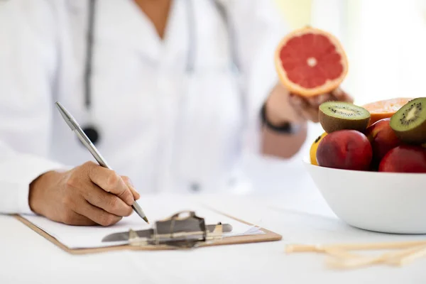 Adult caucasian nutritionist doctor in white coat at table makes notes in clipboard, shows grapefruit in clinic office interior, cropped. Weight loss and health care planning diet