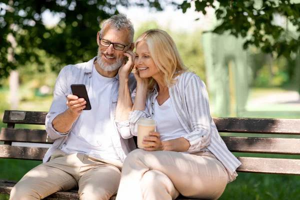 Happy mature couple listening music on smartphone together outdoors in park, smiling senior spouses sharing earphones while relaxing on bench outside, woman drinking takeaway coffee, copy space