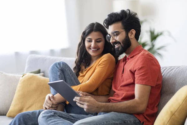 Young Smiling Indian Couple With Digital Tablet Relaxing On Couch At Home, Happy Millennial Eastern Spouses Shopping Online Or Browsing Internet While Resting On Sofa In Living Room, Free Space