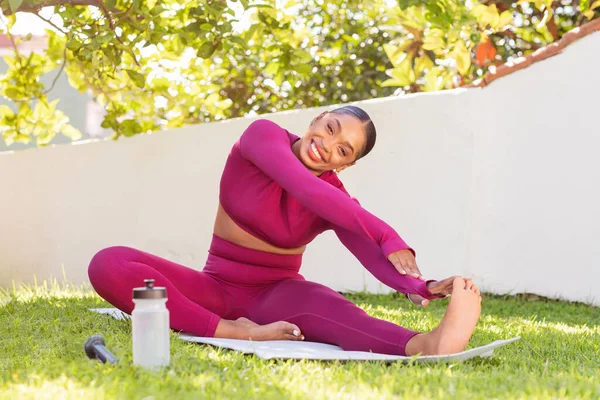 Athletic smiling well-fit attractive young black woman wearing pink sportswear exercising on fitness mat at garden, doing stretching yoga pilates, copy space. Outdoor workout