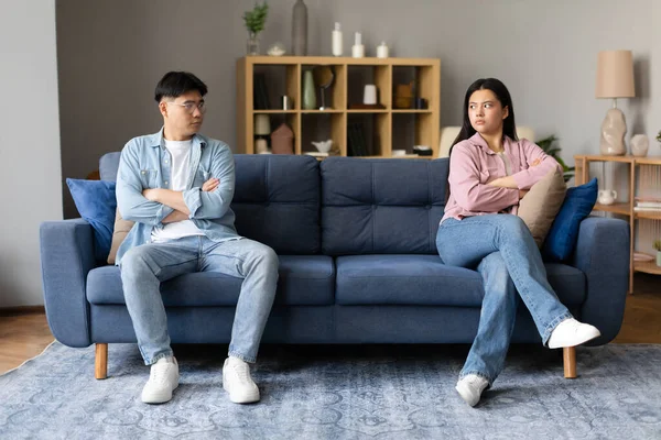 Family Conflicts, Breakup. Unhappy Offended Japanese Young Couple Sulking Sitting On Sofa, Looking At Each Other With Discontent, Having Issue With Communication At Home. Relationship Problems