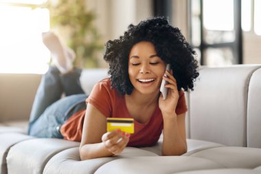 Evde kredi kartı tutan ve cep telefonuyla konuşan gülümseyen genç siyahi kadın, evinde rahat bir koltukta yatarken internetten alışveriş yapan mutlu Afrikalı Amerikalı kadın,
