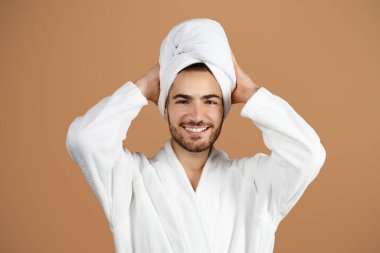 Sağlık ve Güzellik. Arkasında el ele tutuşan, kafaya dokunan, beyaz bornoz giyen, bej rengi stüdyonun arka planında rahatlayan ve spa günü yapan Latin bir adamın portresi.