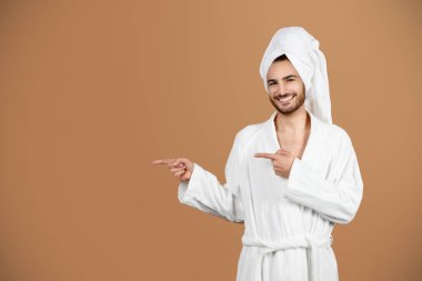 Erkek Güzellik Teklifi. Fotokopi uzayını işaret eden mutlu Latin genç adam, şımartma ve kişisel bakım için kozmetik reklamları yapan, beyaz bornoz giyen ve kahverengi arka planda havluyla gezen