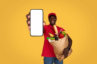 Bin yıllık Afro-Amerikan kurye adam elinde yiyecek dolu kağıt torba boş ekranlı telefon, turuncu stüdyo arka planında izole edilmiş. Reklam, teklif, eve sipariş verme uygulaması, servis