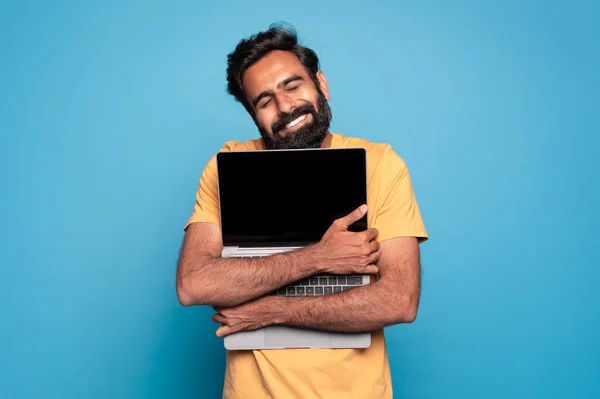 兴奋的印度男人抱着笔记本电脑 用黑色的空白屏幕 紧紧抓住笔记本电脑的胸部 微笑着 男性科技爱好者在蓝色的工作室背景下摆出孤立的姿势 复制空间 — 图库照片