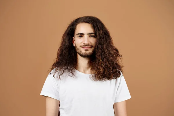 Portrait Of Handsome Latin Guy With Long Curly Hair Wearing T-Shirt Standing On Brown Beige Studio Background. Confident Young Man In Casual Posing Looking At Camera Advertising Your Offer