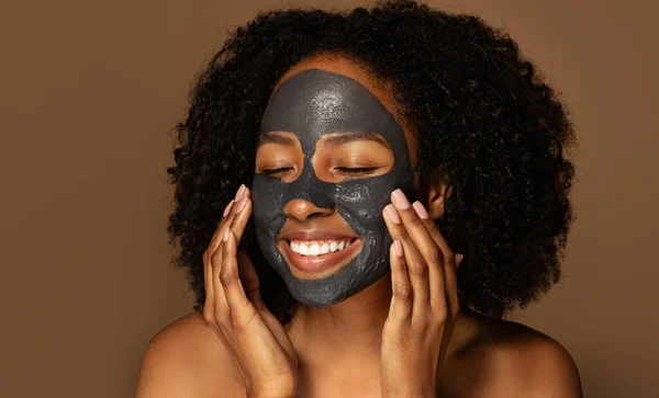 Closeup of smiling attractive millennial topless african american lady using black mask, isolated on brown background. Peel-off face mask for deep skin cleanse, face care routine concept