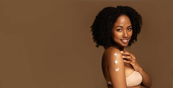 Happy pretty half-naked young black woman with smooth youth-looking skin apply hydrating, moisturizing beauty product cream on shoulder, smiling, brown background, copy space. Skin care routine