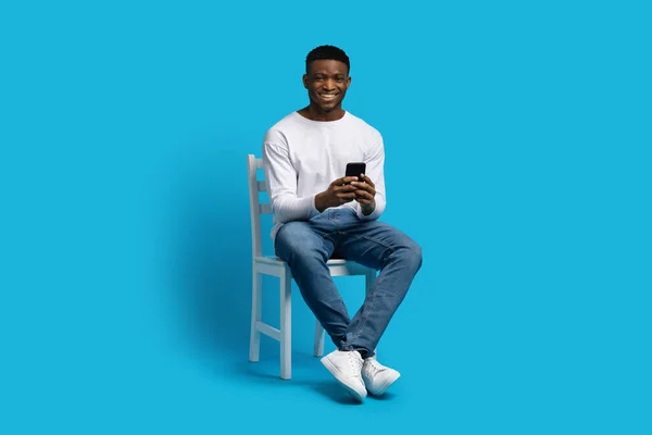 ポジティブなハンサムな若いアフリカ系アメリカ人男性が椅子に座り スマートフォンを使って微笑み 青い背景に隔離され スペースをコピーします モバイルアプリのデート — ストック写真