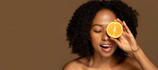 Vitamin C in cosmetology, skin whitening. Hot half-naked young black woman holding orange over one eye for rejuvenation, skin hydration, beauty treatment isolated on brown background, copy space