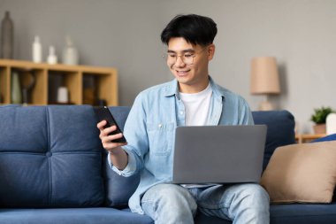 İnternet ve Gadget 'lar. Gülümseyen Japon adam akıllı telefon kullanıyor dizüstü bilgisayarla oturuyor, evdeki koltukta iletişim kuruyor ve çalışıyor, oturma odasındaki telefonda iş uygulamasını kullanıyor.