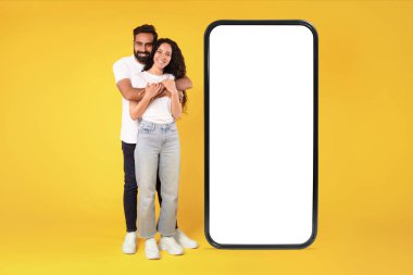 Neşeli Arap çift, sarı arka planda beyaz ekranlı akıllı telefonun yanında durup sarılıyorlar. Cep telefonu uygulaması ya da iletişim reklamı için model alıyorlar. Tam boy çekim