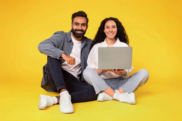 笑顔の中東カップル ラップトップコンピュータを使用して 床に座って黄色いスタジオの背景でカメラを見ます 男性と女性ウェブサーフィンウェブサイトとコンピュータ上でオンライン映画を見る — ストック写真