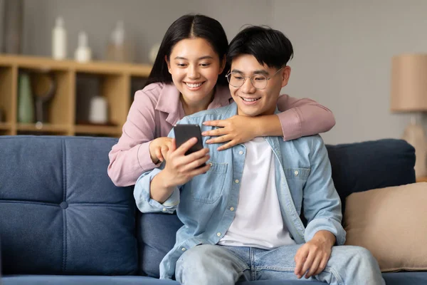 Cheerful korean couple sharing social media on mobile phone, happy asian family sitting on sofa, using smartphone gadget at home interior, woman embracing her husband looking at cellphone screen